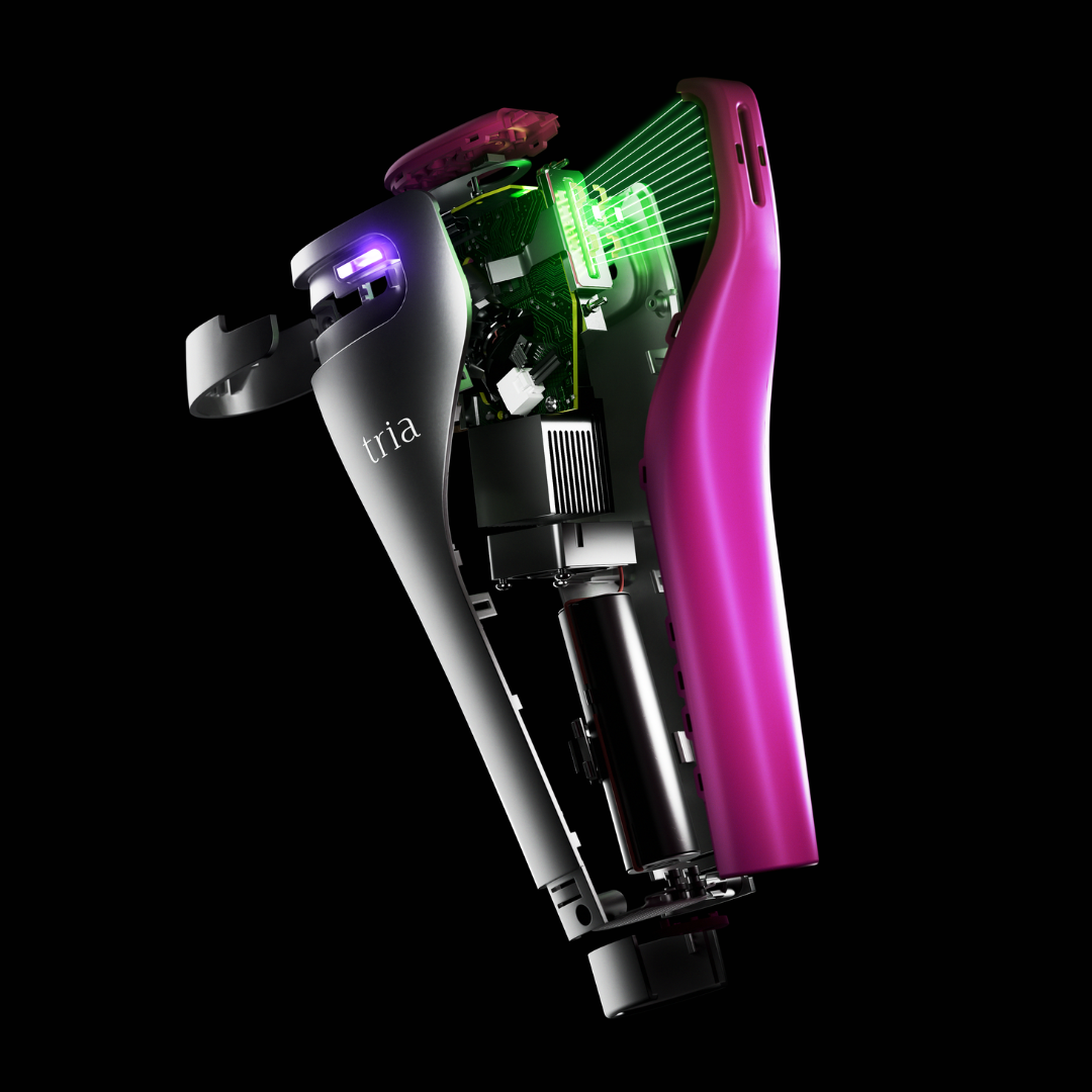 A Tria FRX Skin Rejuvenation Laser shown at an angle on a solid black background, with its outer shell removed to reveal internal components. The device has a black exterior with a prominent bright pink inner sleeve, green illuminated internal elements, and a purple light near the top.